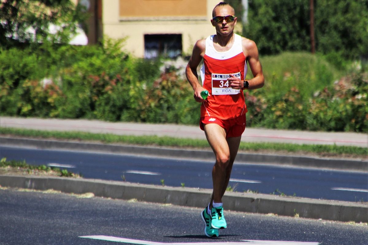 zdjęcie przedstawia biegnącego mężczyznę, ubranego na sportowo z numerem, w tle ulica, zieleń, budynki.